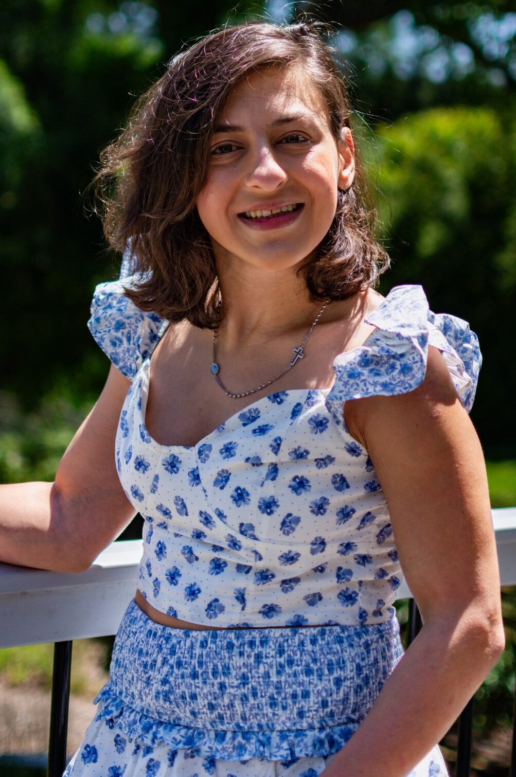 Yasmina Saba smiling outdoors, standing by a railing in a blue and white floral dress with trees and greenery in the background.